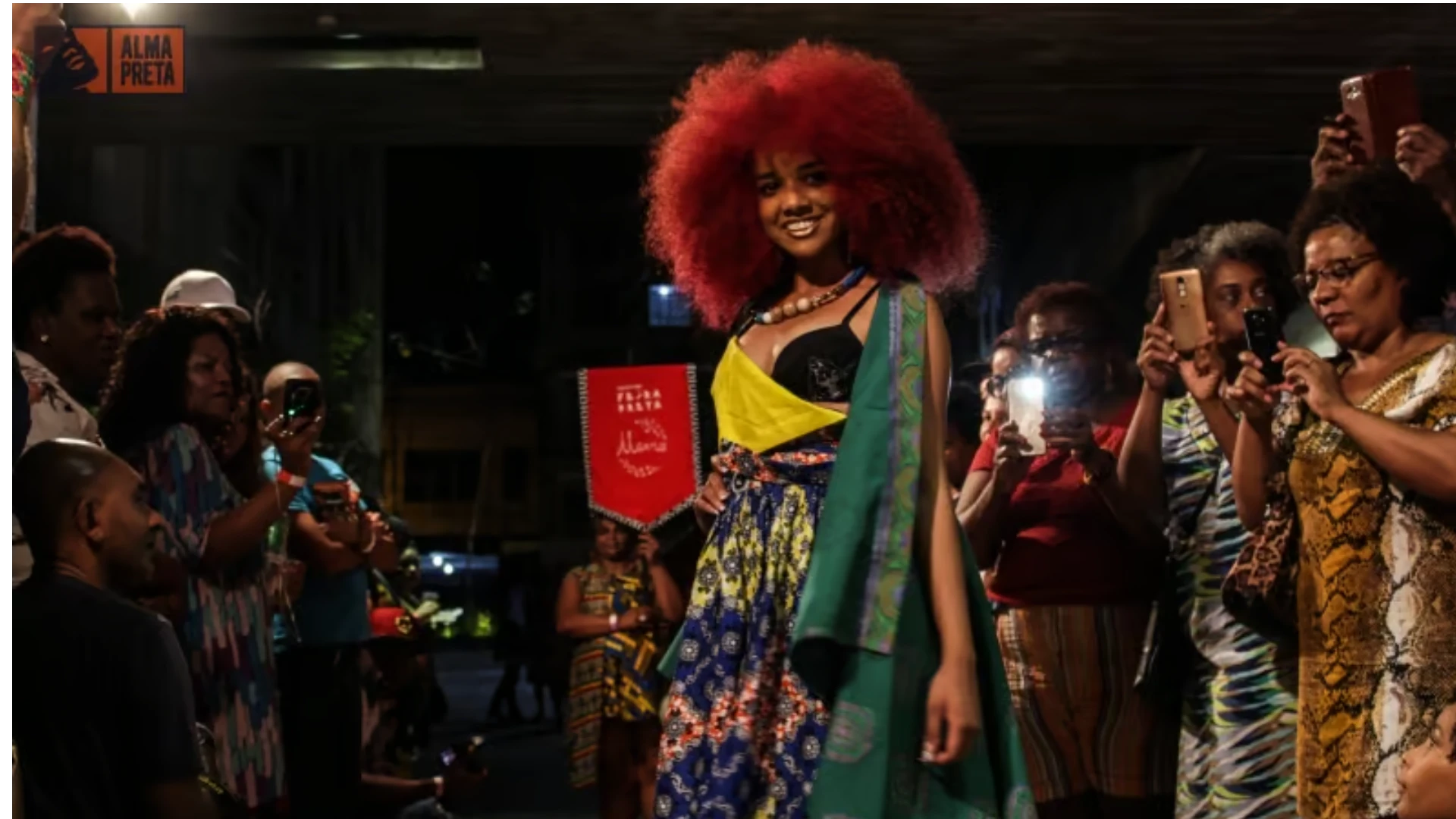 Mercado de eventos para empreendedores negros: desfile cultural com mulher negra de cabelo afro vibrante, cercada por público engajado.