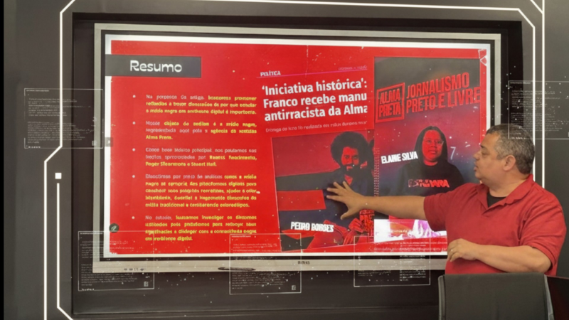 Um homem negro de camisa polo vermelha, de meia-idade, está em pé na frente de uma grande tela. Ele aponta para o lado direito da tela, onde há uma imagem de uma revista com o título "ALMA JORNALISMO PRETO E LIVRE". O lado esquerdo da tela mostra texto em português em um fundo vermelho. Há reflexos e algumas barras digitais transparentes sobrepostas na tela.